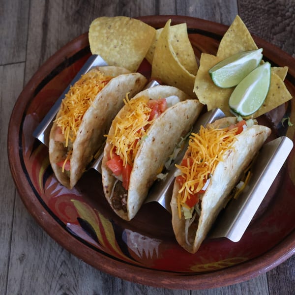 Three tacos with cheese, tomatoes, and lettuce, served with tortilla chips and lime wedges on a decorative plate.