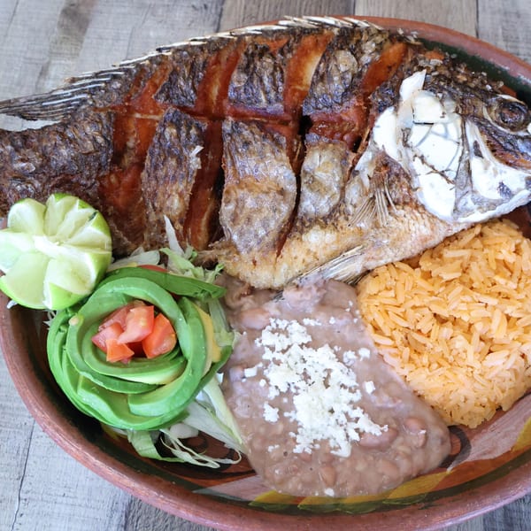 Fried whole fish with rice, refried beans, avocado, and lime on a rustic plate.