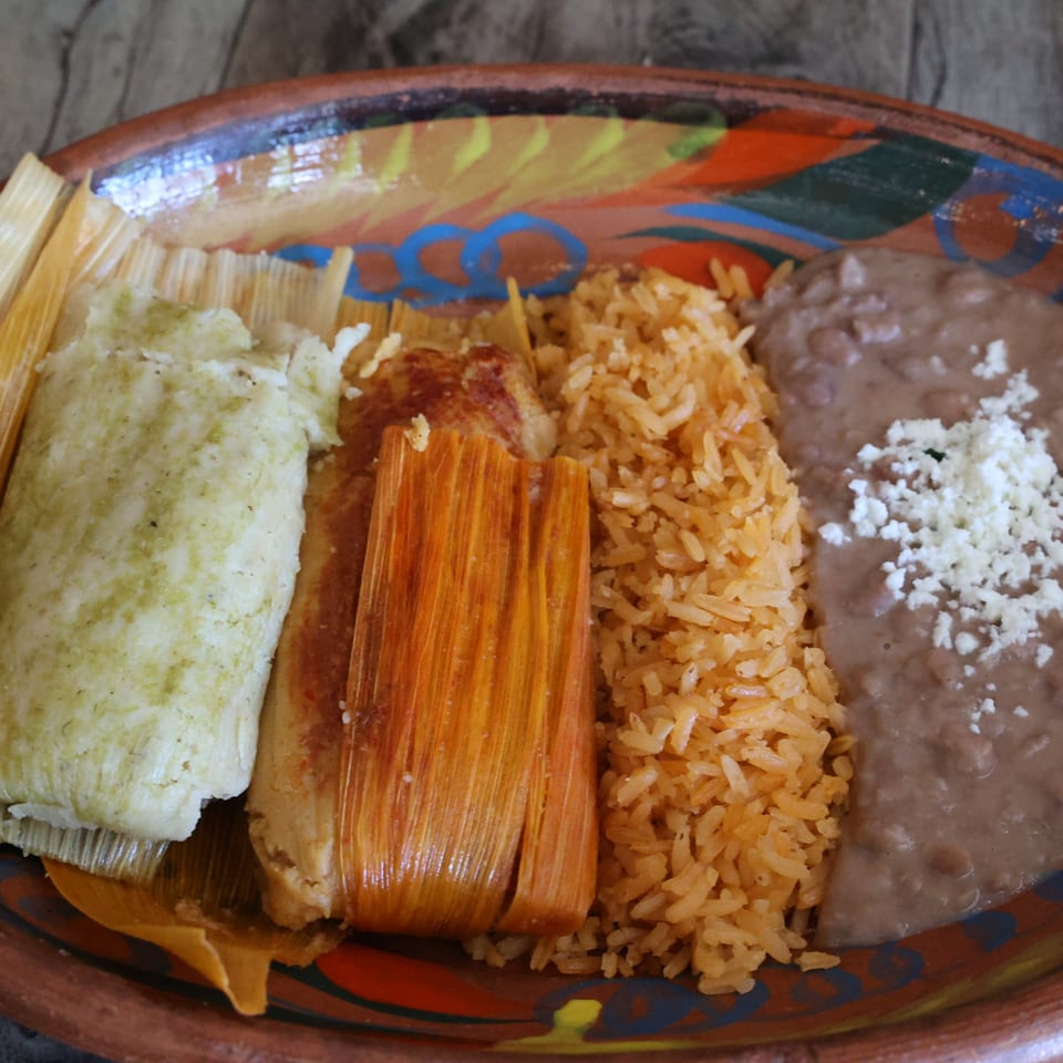 A colorful plate of Mexican tamales, rice, and refried beans with crumbled cheese.