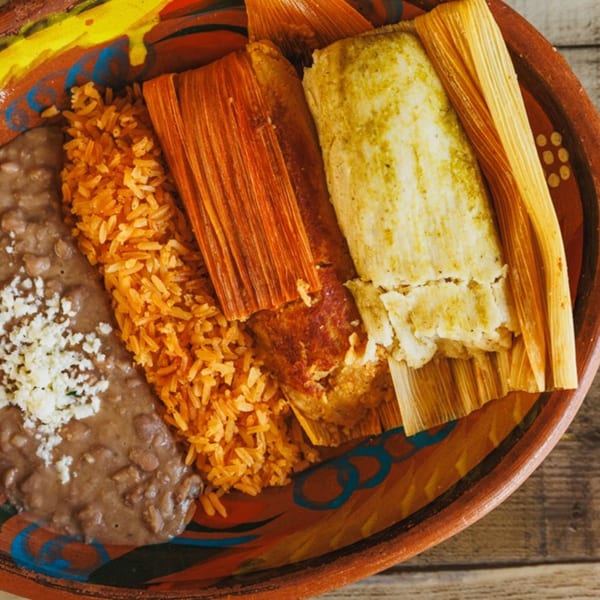 Two tamales, one red and one green, with rice and refried beans in a colorful bowl.