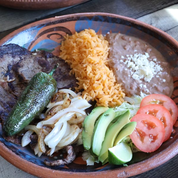 Mexican dish with steak, grilled pepper, onions, rice, refried beans, avocado, tomatoes, and lime.